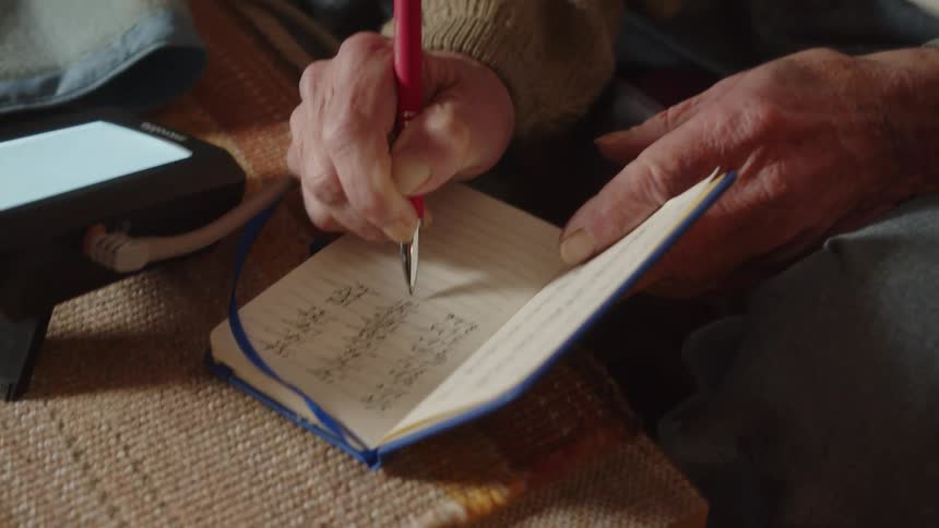 A man is sitting on his living room couch looking at the small screen on a machine as he writes in a notebook