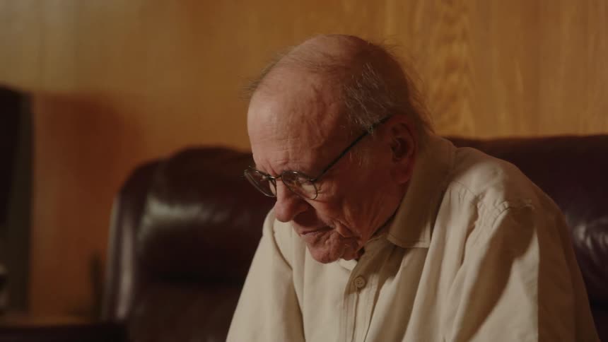 An older man wearing glasses sitting on his couch reaching his arm out and then resting them on his knees