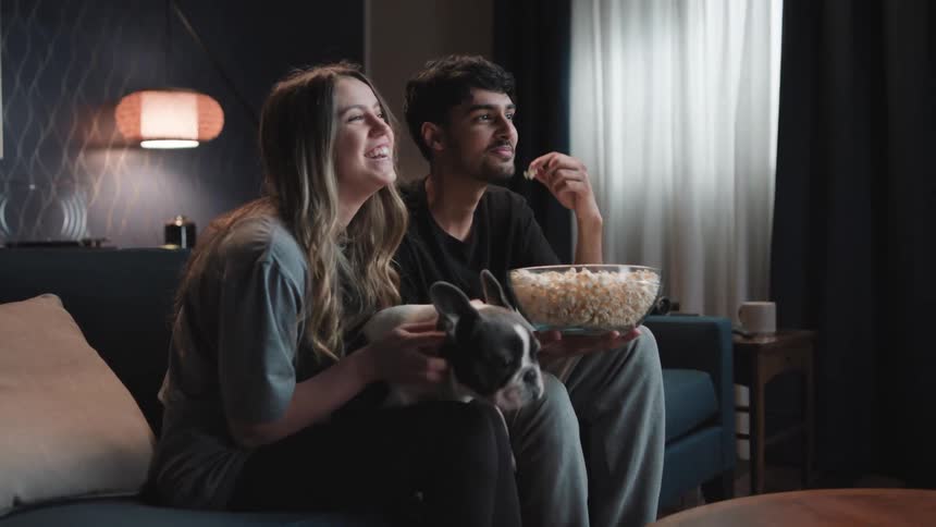 Man and woman sitting on couch eat popcorn and pet dog in the living room