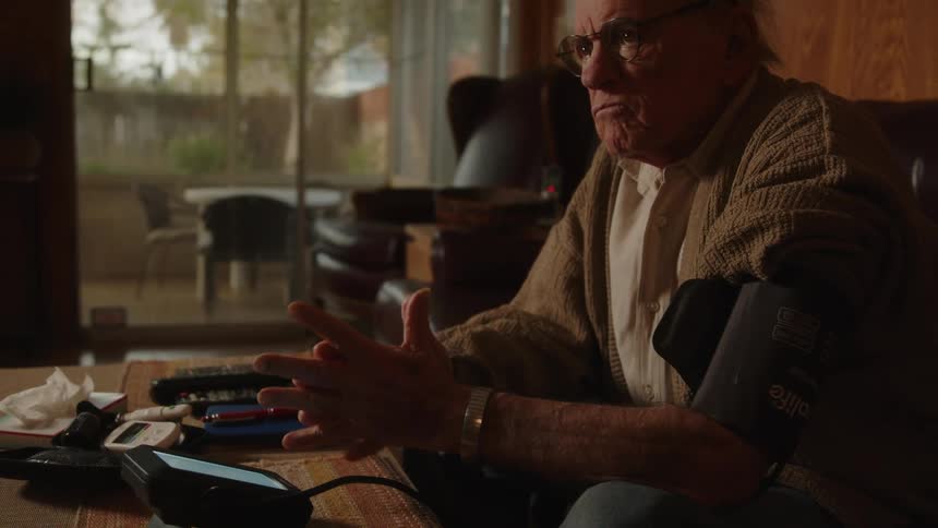An older man wearing glasses and a blood pressure cuff on his arm sitting on the edge of his couch rubbing his hands together