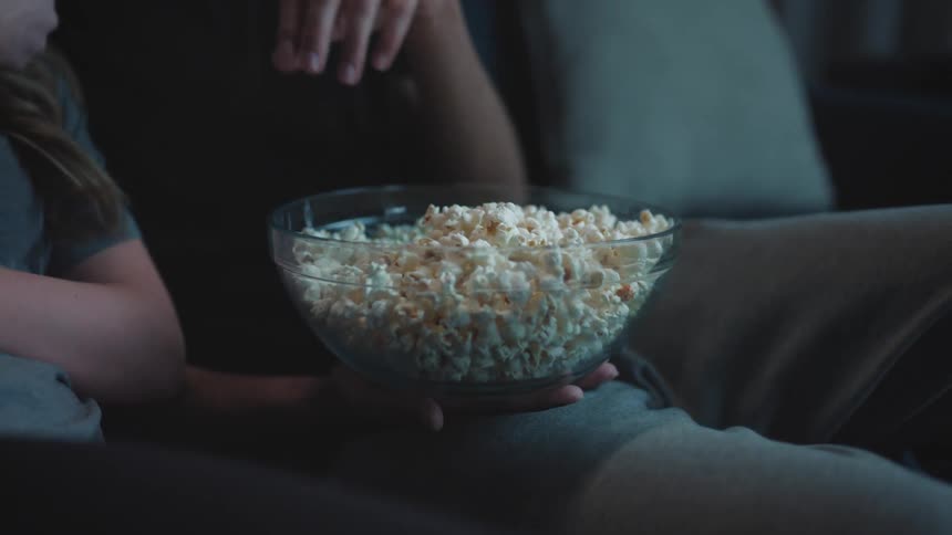 Two people are sharing a bowl of popcorn and taking turns grabbing popcorn while the lighting from a screen is reflecting on the couch they are sitting on