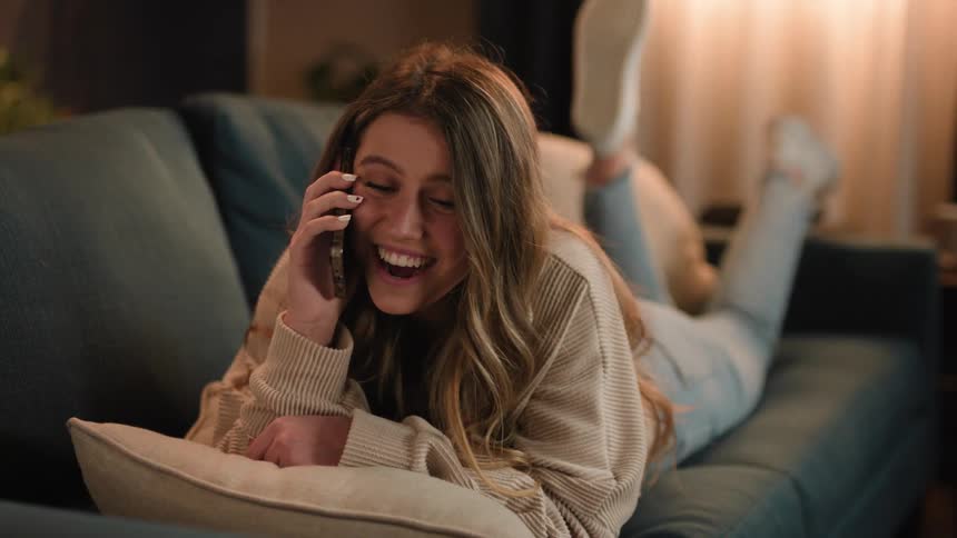 A teenage girl lies on a couch at home and kicks her feet as she talks on a cell phone and laughs