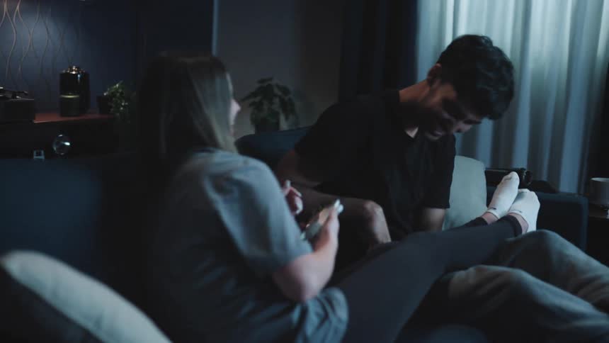 Woman sitting on couch with man takes his picture on her smartphone then they take a selfie together