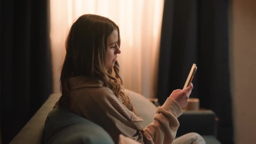 Young woman sitting at the couch at home scrolls on her smartphone
