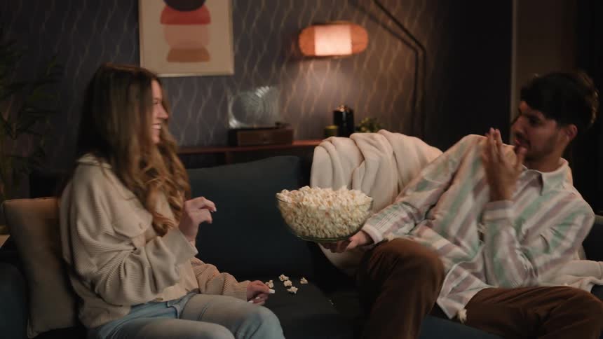Young couple sitting on a couch at home plays with popcorn in the living room