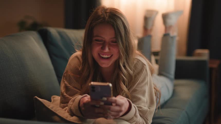 Beautiful young woman laying on the couch at home smiles while on a video call on her phone