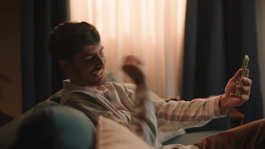 Young man sitting on the couch at home haves fun on a video call through his smartphone