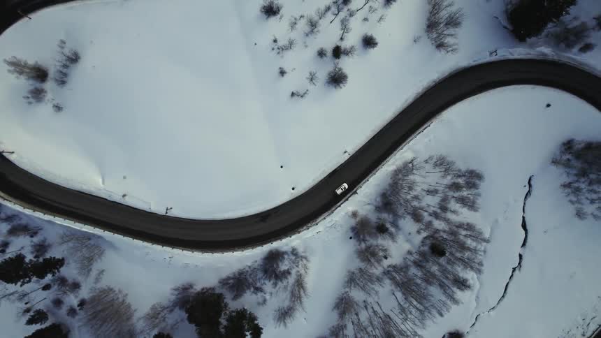 Top down aerial view of a white vehicle driving on a road through the snow covered landscape