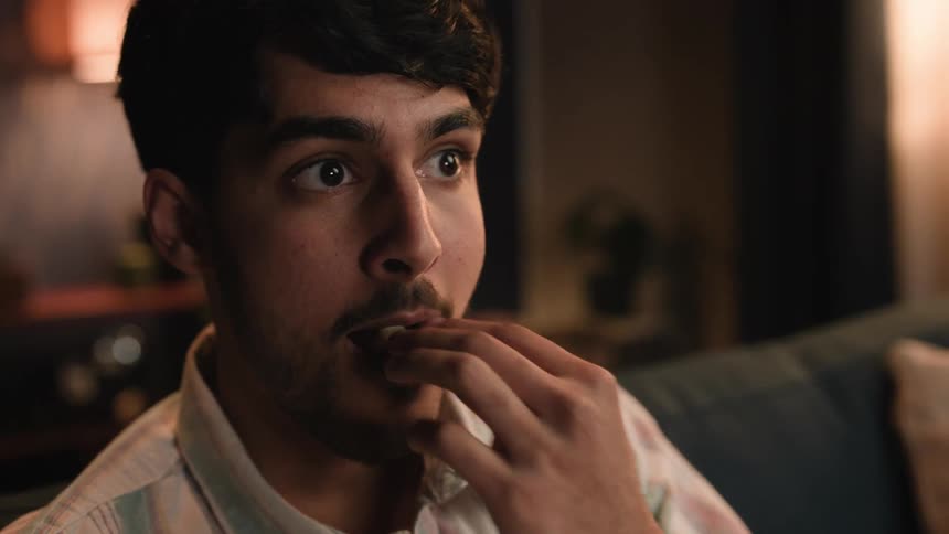 A man seems to be watching a movie eating popcorn while scared of what he is watching and then relieved
