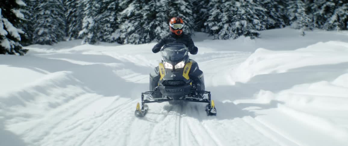 A person rides a snowmobile with headlights on on a road through the pine tree forest at daytime