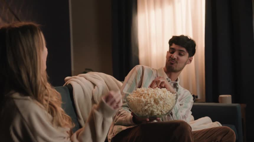 A man is sitting on a couch holding a bowl of popcorn with a young woman as they throw popcorn into each others mouths