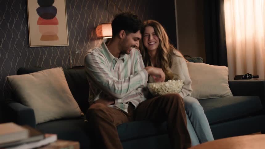 A man sitting on a couch when a woman sits down next to him grabbing the bowl of popcorn and throwing it in each others mouths