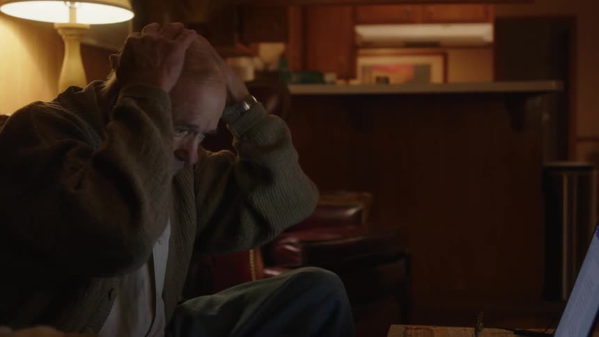 An elderly man sitting in front of his laptop contemplates and rubs his head in distress