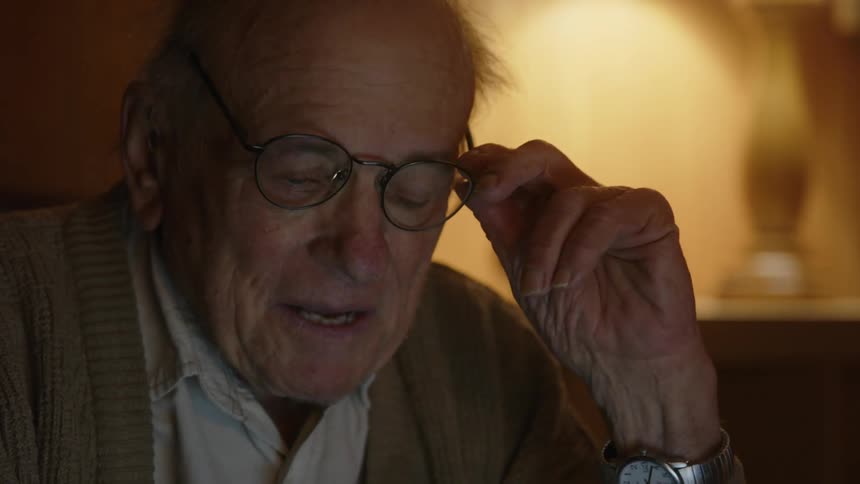 An elderly man vigorously works on something while sitting and smiles to himself