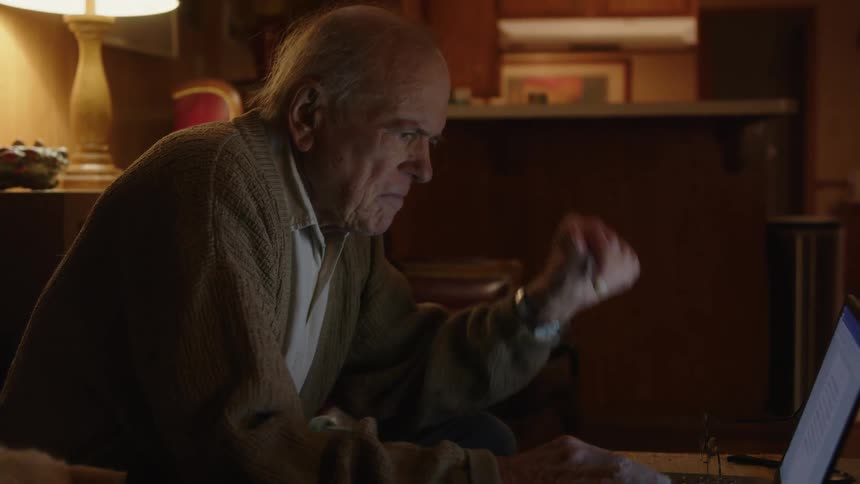An elderly man at a desk in a room types on a laptop