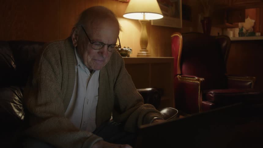Elderly man sitting on a couch at home puts his glasses on to type on a laptop over the coffee table