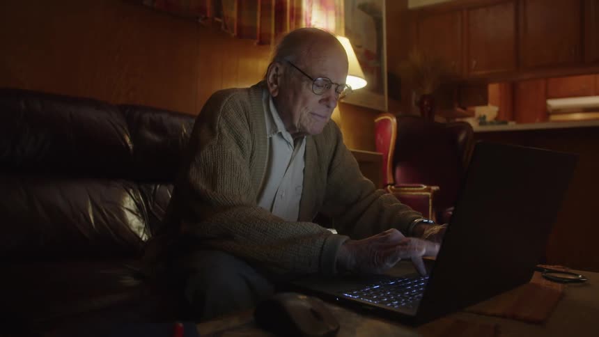 An older man sitting on the edge of his couch typing on his laptop in front of him