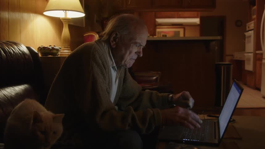 A man is typing on his laptop at home with his cat sitting nearby
