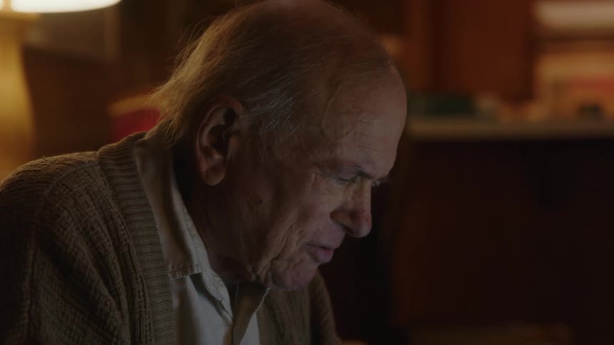 An older elderly man is sitting down inside of his home