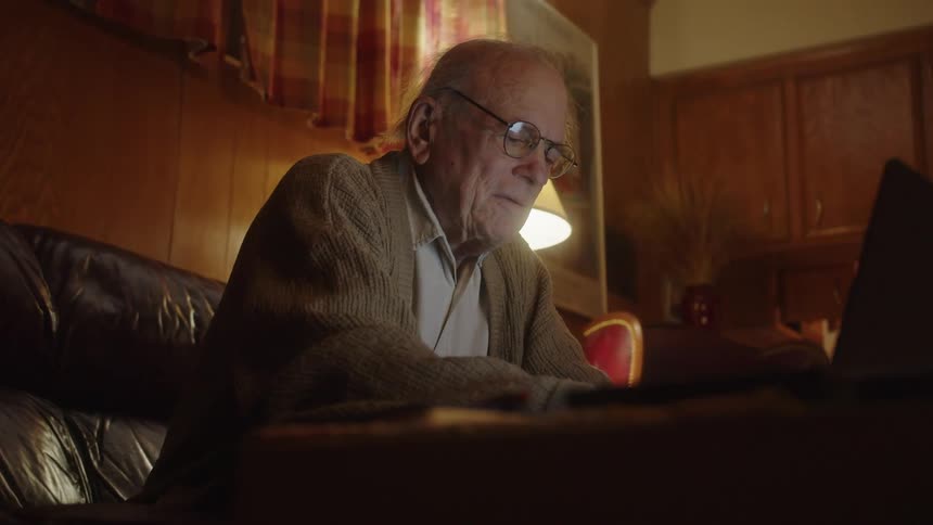 An elderly man types on a laptop while seated at a desk and smiles to himself
