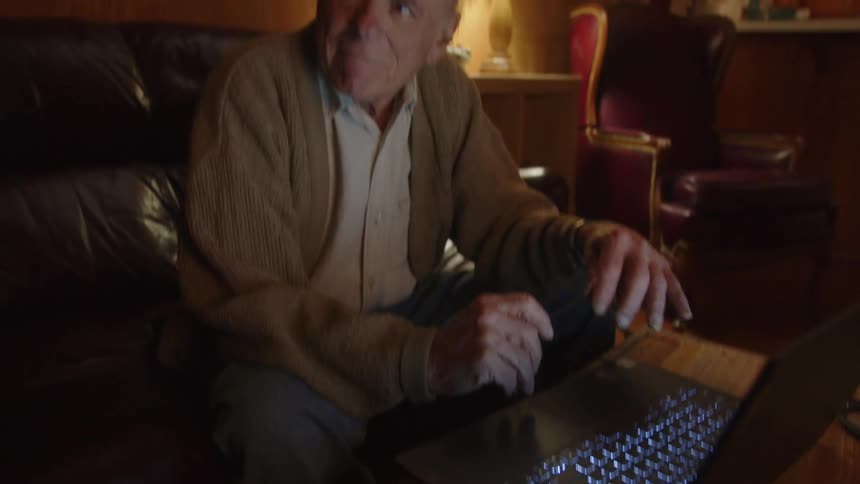 An elderly man sitting in front of his laptop looks to the side and smiles while gesturing