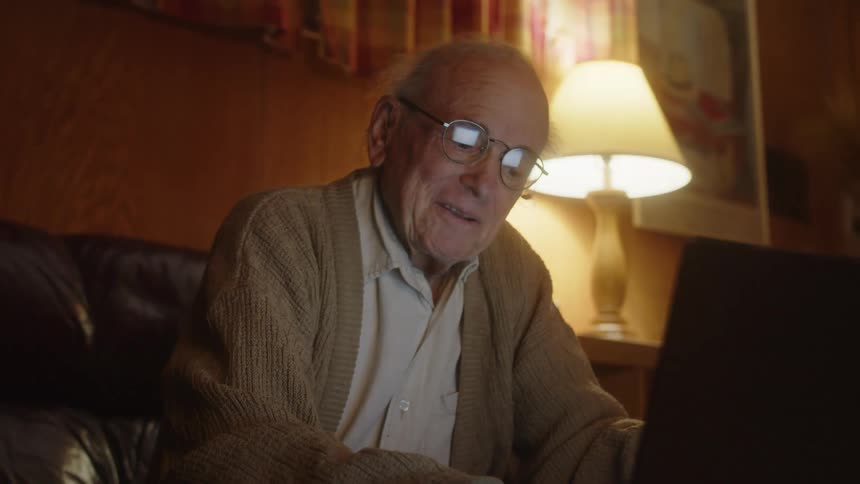 A man is sitting in his living room smiling and laughing as he has a conversation on his laptop computer