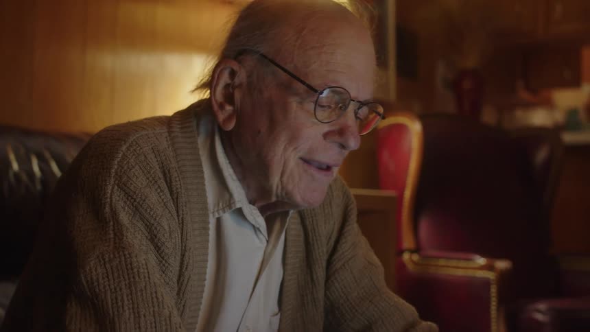 An elderly man sitting on his couch types on his laptop while looking amused
