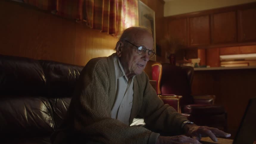 An older elderly man is sitting down and on his laptop