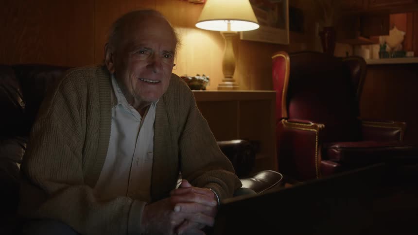 Elderly man with brown jacket and shirt watches television laughing and typing on laptop in the living room at night