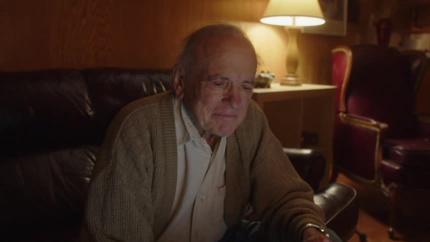 An elderly man sits at home and types on a laptop and smiles and looks up as he thinks