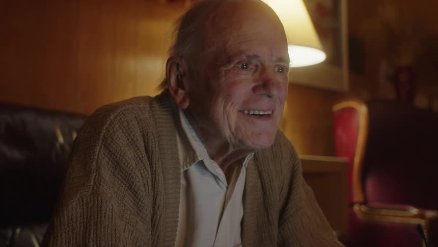 An older man sitting on the edge of his couch in front of a laptop smiling and laughing and then putting his glasses on