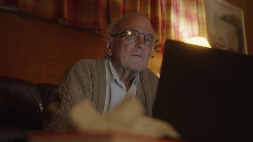 Elderly man sitting on a couch at home thinking while typing on a laptop over the coffee table