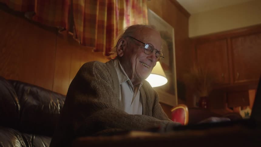 An older man wearing glasses sitting on the edge of his couch typing on the laptop in front of him