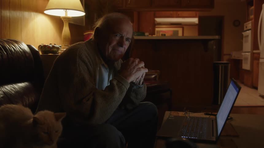An older man sitting on the edge of his couch with a cat next to him and a laptop in front of him