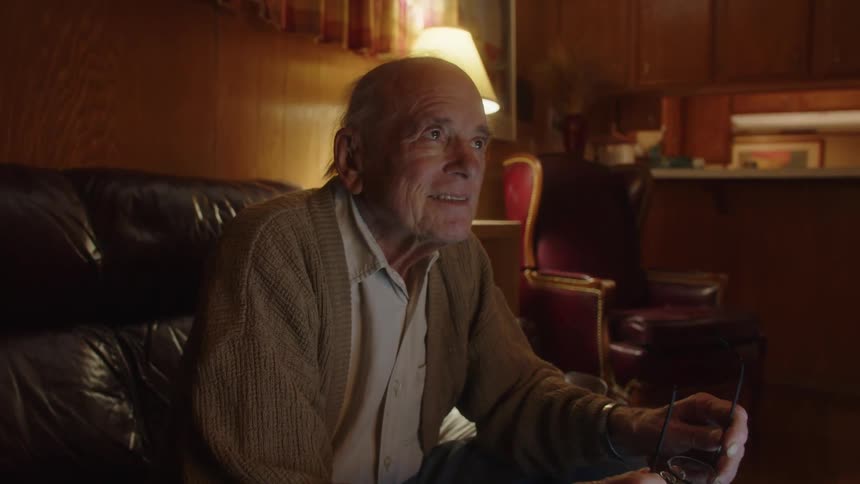 An elderly man sitting on his couch in the living room looks around in thought and starts working at his laptop