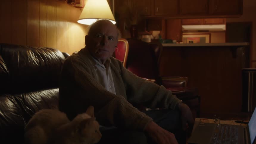 An elderly man sitting on a couch looks at his laptop exasperated and thinking