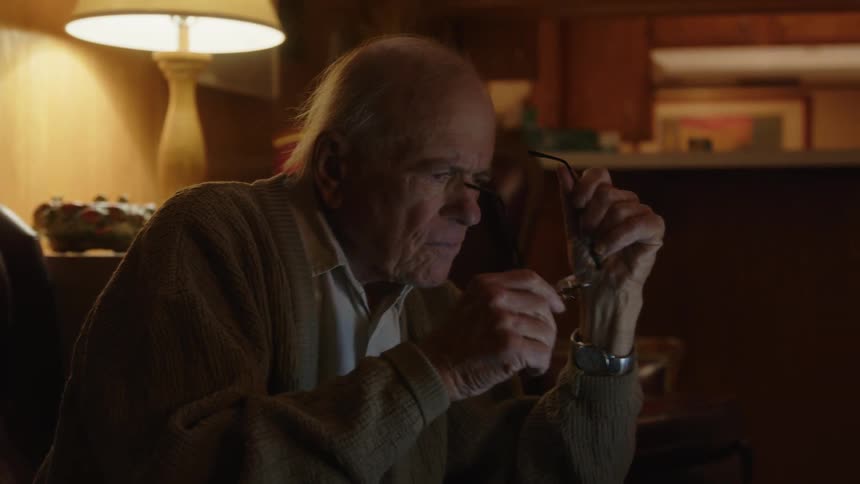 Elderly man sitting on a couch at home puts his glasses on to read on a laptop over the coffee table