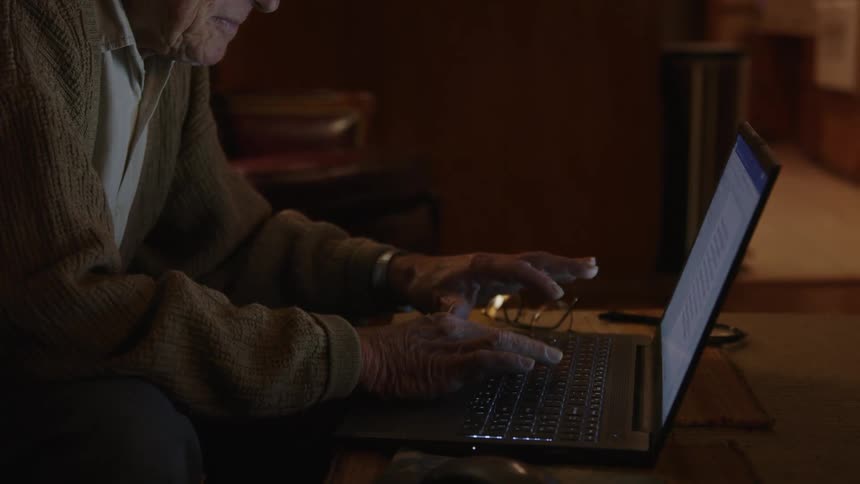 An elderly man at a desk indoors types on a laptop and puts on his glasses
