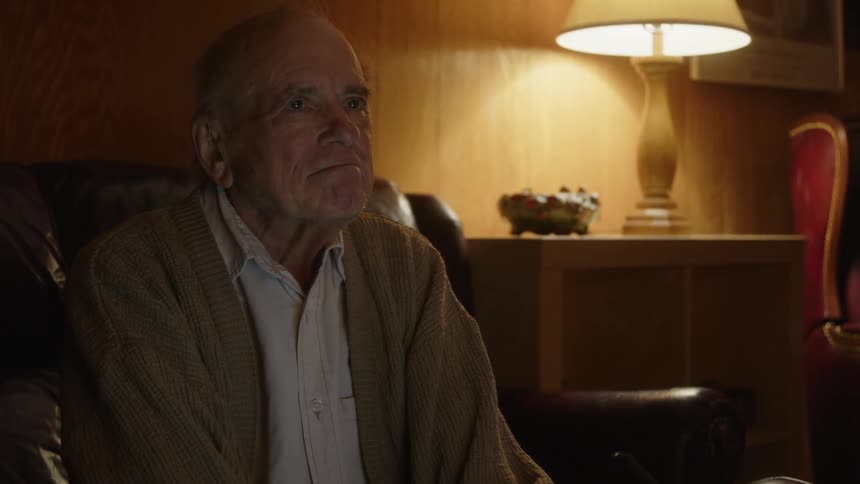 An older elderly man is sitting down inside of his home