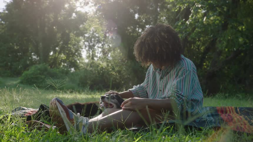 Woman with curly hair and blue shirt sits on lawn petting dog in the field