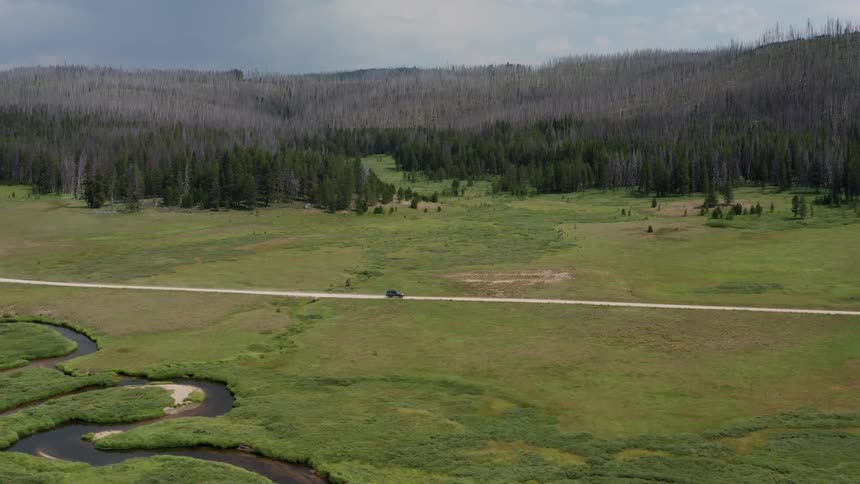 An aerial view of a vehicle driving down a road in a grassland past a river and forest