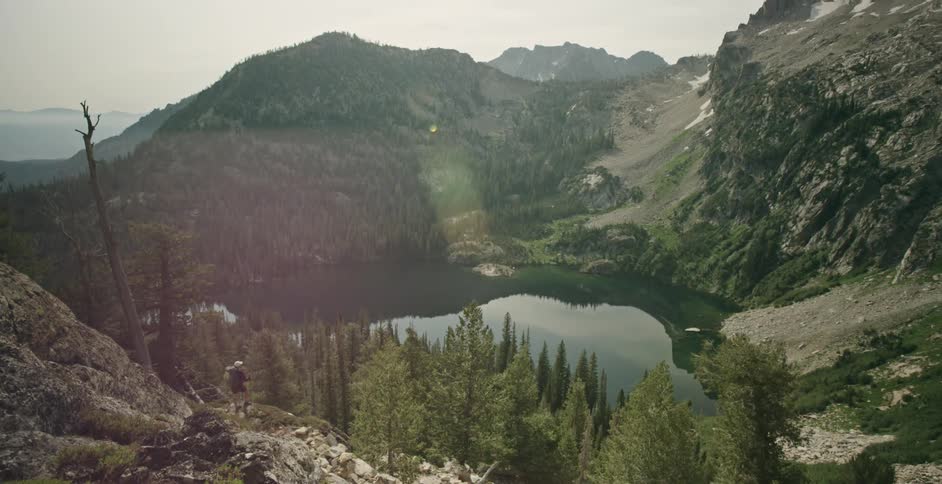 A hiker stands on a mountainside looking at the lake in a forested mountain valley