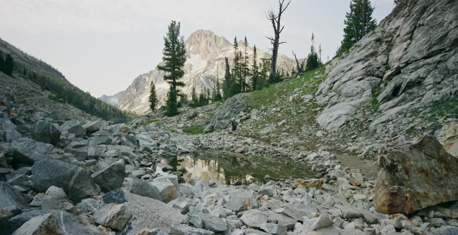 A person hiking on a mountain trail passes by a small body of water surrounded by loose rocks