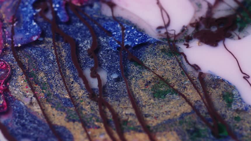 Close up of brown liquid ink pouring over a chaotic mix of colorful inks and glitter