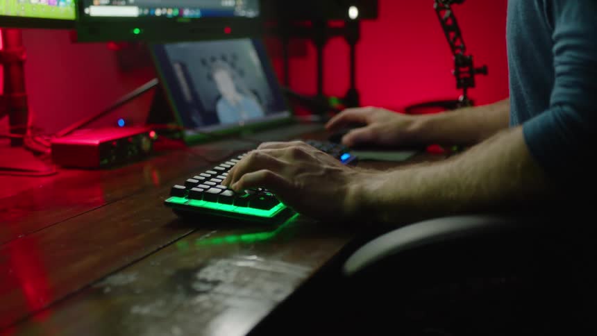 A man is sitting at a desk typing on a keyboard with a tablet computer in front of him and two monitors on the wall