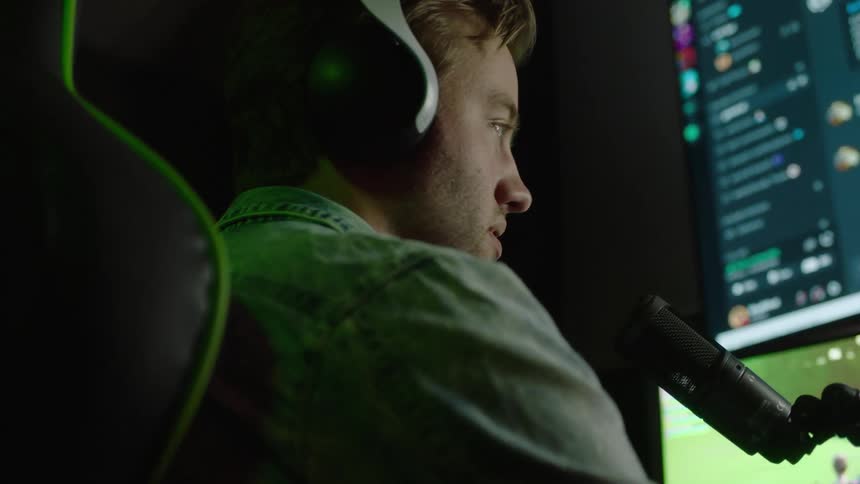An up close of a man sitting at a computer with headphones and a microphone while playing a game on a laptop