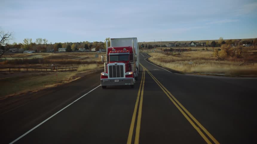 A semitruck is traveling down a highway in a rural area during the autumn