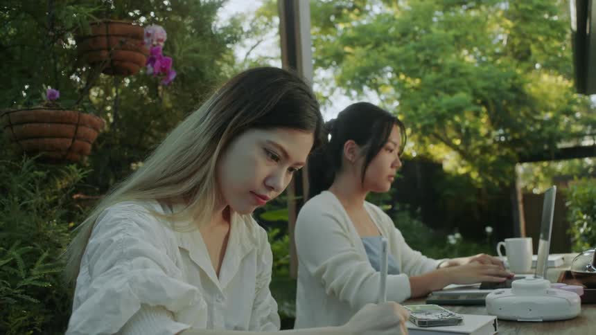 Two women sitting at a table outdoors one typing on her laptop and the other writing on paper