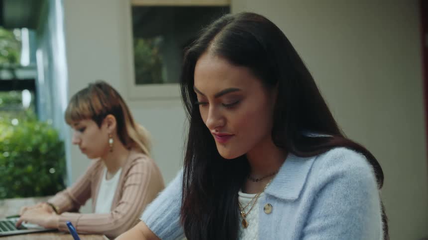 Two women sit outside at a table one typing on a laptop and the other writing with a pen