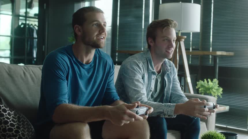 Two men are sitting on the couch with video game controllers in their hands laughing and playing games